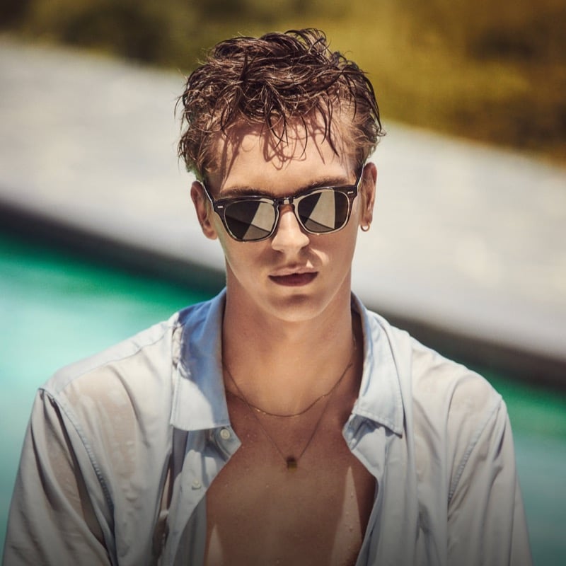 Dominik Sadoch in Oliver Peoples' geometric Arliss sunglasses and an open pale button-up shirt beside a pool, from the brand's fall 2026 Twice Upon a Time story