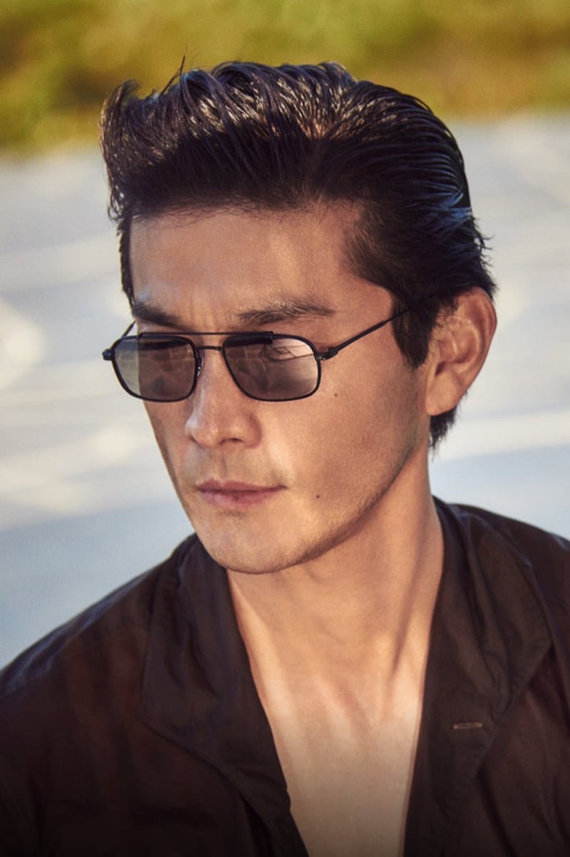 Daisuke Ueda in Oliver Peoples' black rectangular Caylor sunglasses and a dark shirt, backlit by warm evening light, from the brand's fall 2026 Twice Upon a Time story
