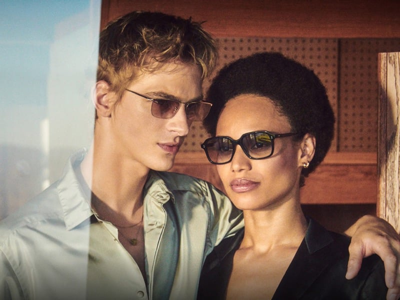 Dominik Sadoch in Oliver Peoples' gold-framed EZ-Y sunglasses rests an arm around Afrodita Dorado, who wears black square Tassin sunglasses, in a wood-paneled room, from the brand's fall 2026 Twice Upon a Time story