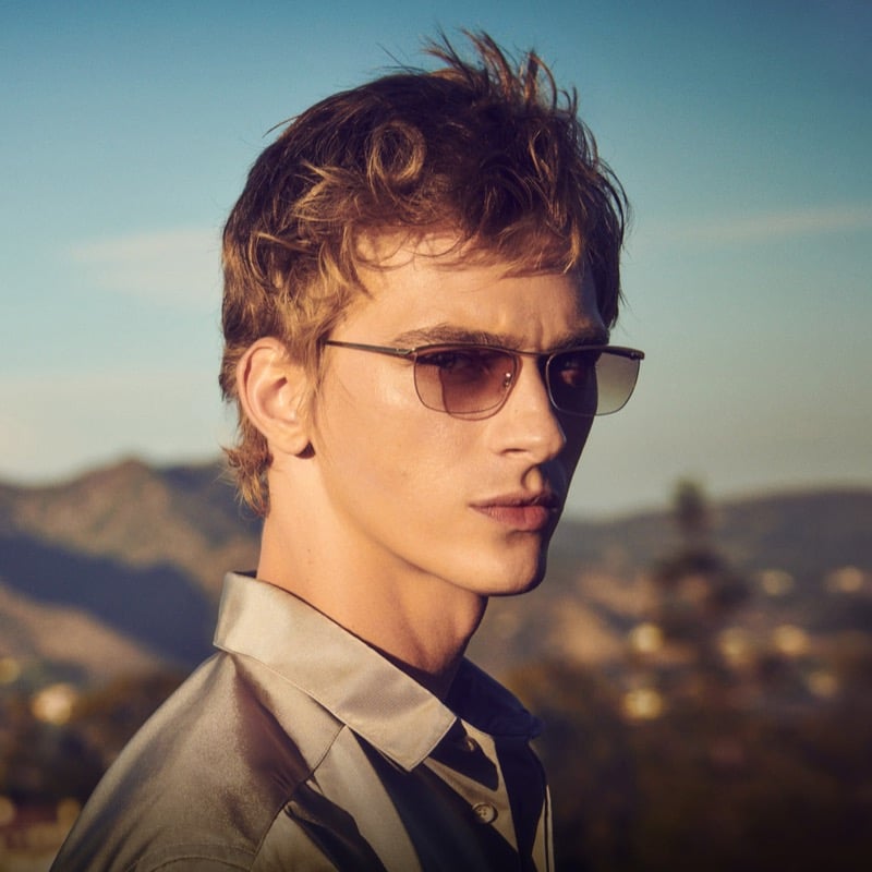 Dominik Sadoch in Oliver Peoples' thin-metal Ez-Y sunglasses and an olive-toned satin shirt against a hillside backdrop, from the brand's fall 2026 Twice Upon a Time story