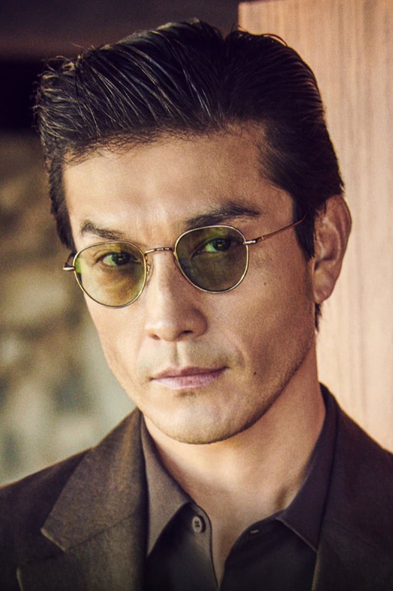 Daisuke Ueda in Oliver Peoples' round, gold-rimmed TK-16 sunglasses with green-tinted lenses and a brown blazer, from the brand's fall 2026 Twice Upon a Time story