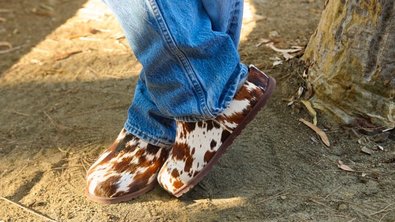 Orville Peck's feet in cow-print UGG clogs on the dirt ground beneath cuffed denim jeans