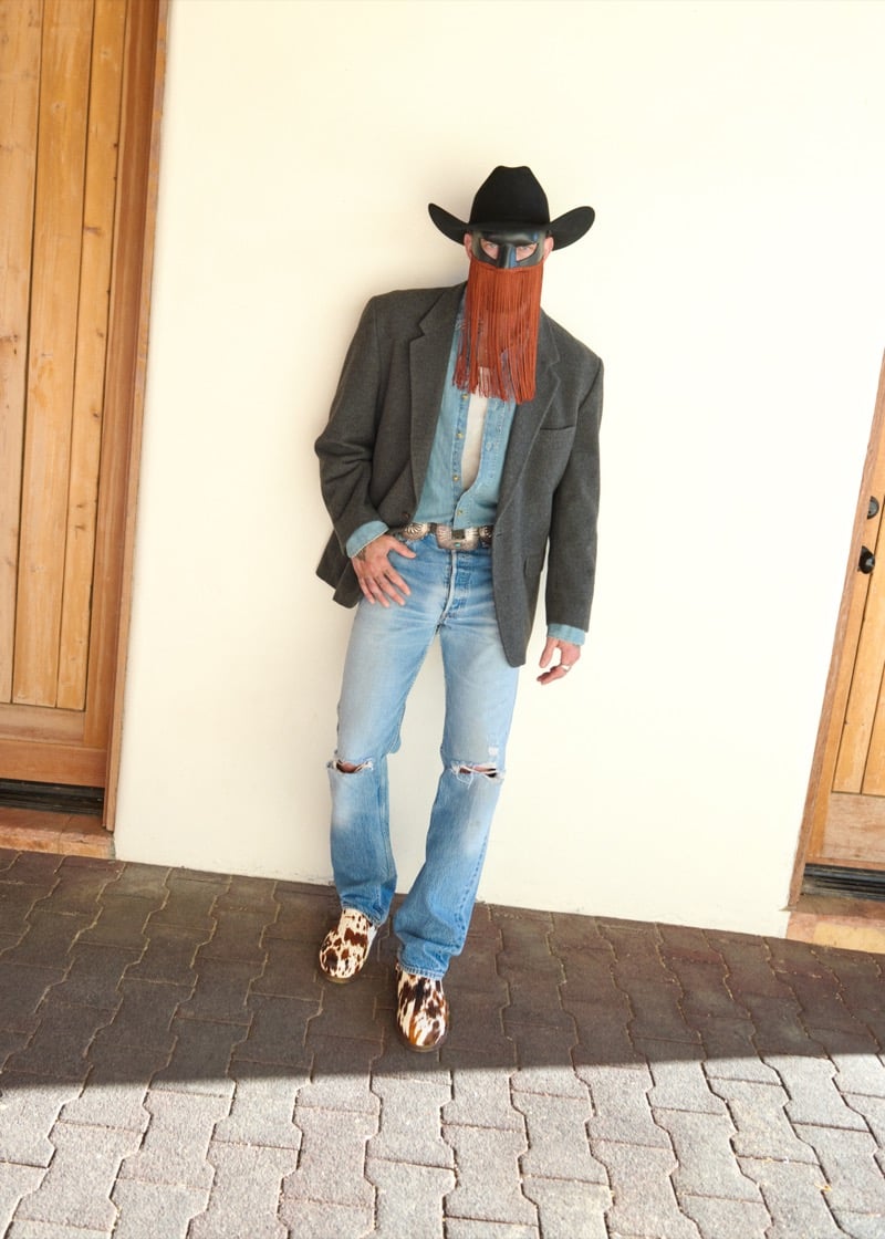 Orville Peck wears a gray blazer over a chambray shirt, an ornate belt buckle, distressed jeans, a black cowboy hat, and cow-print UGG clogs, for UGG's Western collection campaign