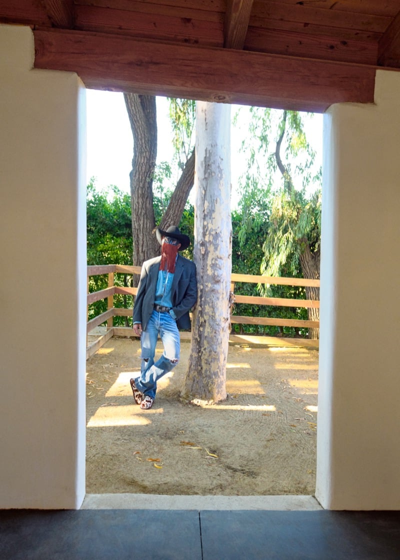 Orville Peck leans against a tree framed by a doorway, wearing a gray blazer, a chambray shirt, distressed jeans, a black cowboy hat, and cow-print UGG clogs, for UGG's Western collection campaign