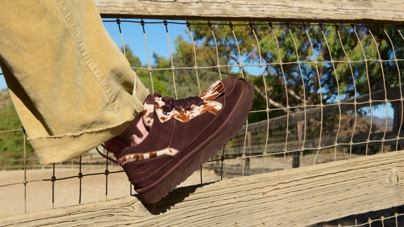 Close-up of a brown suede UGG Western sneaker with a cow-print collar, propped on a wooden fence rail beneath mustard corduroy trousers