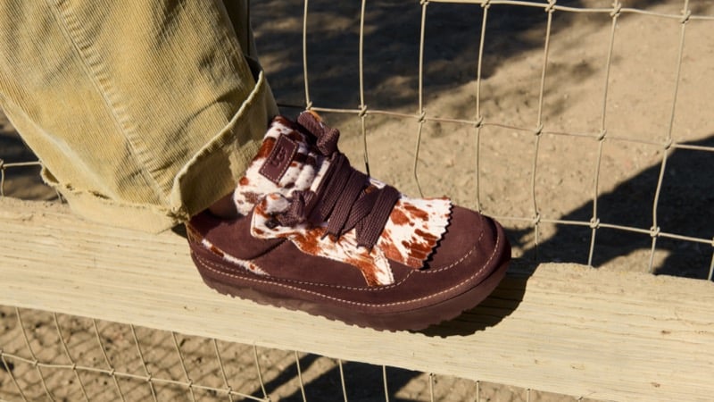 Close-up of a brown suede UGG Western sneaker with a cow-print tongue and laces, resting on a wooden fence rail beneath mustard corduroy trousers