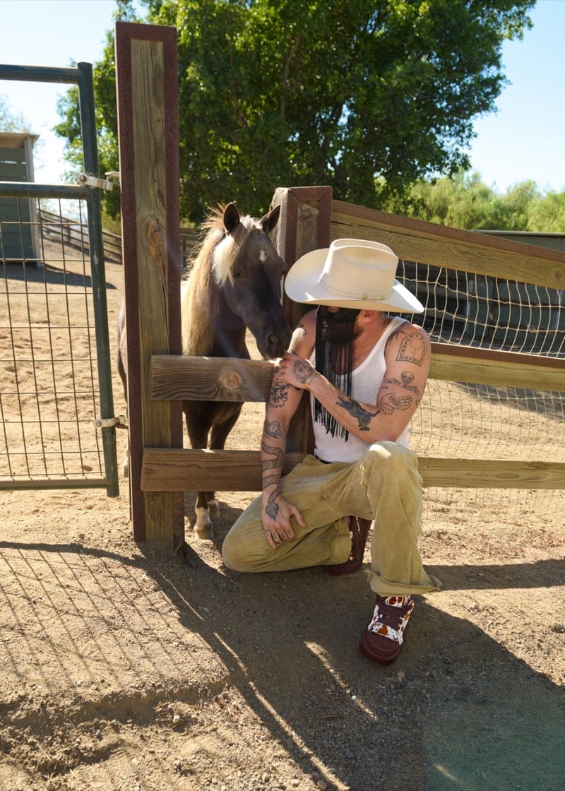 Orville Peck kneels beside a horse at a wooden gate, his tattooed arms bared in a tank top, wearing a wide-brim hat, mustard corduroy pants, and cow-print UGG sneakers, for UGG's Western collection campaign