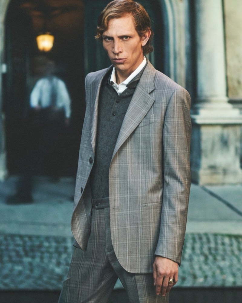 Mike Vasilakis wears a gray checked suit while walking past a lit doorway on a city street for Pal Zileri's fall-winter 2026 campaign.