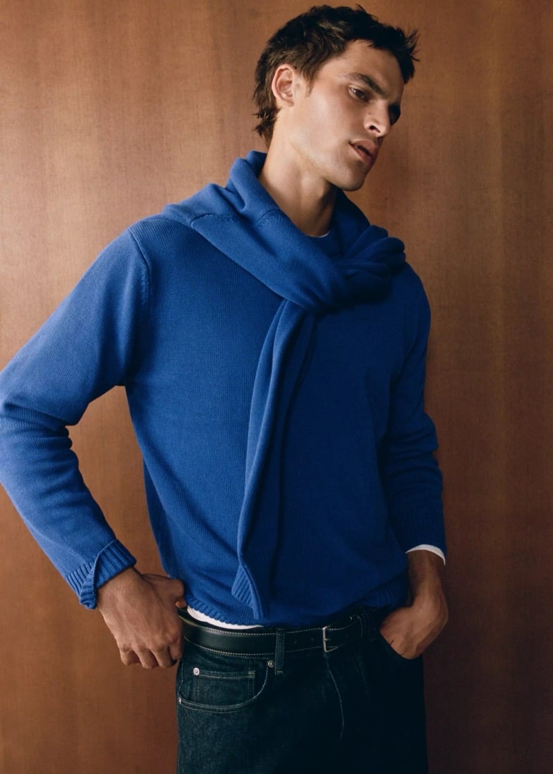 Parker van Noord in a bright blue cotton sweater with a second sweater knotted around his shoulders, wearing dark denim and a stitched leather belt against a wood-paneled wall