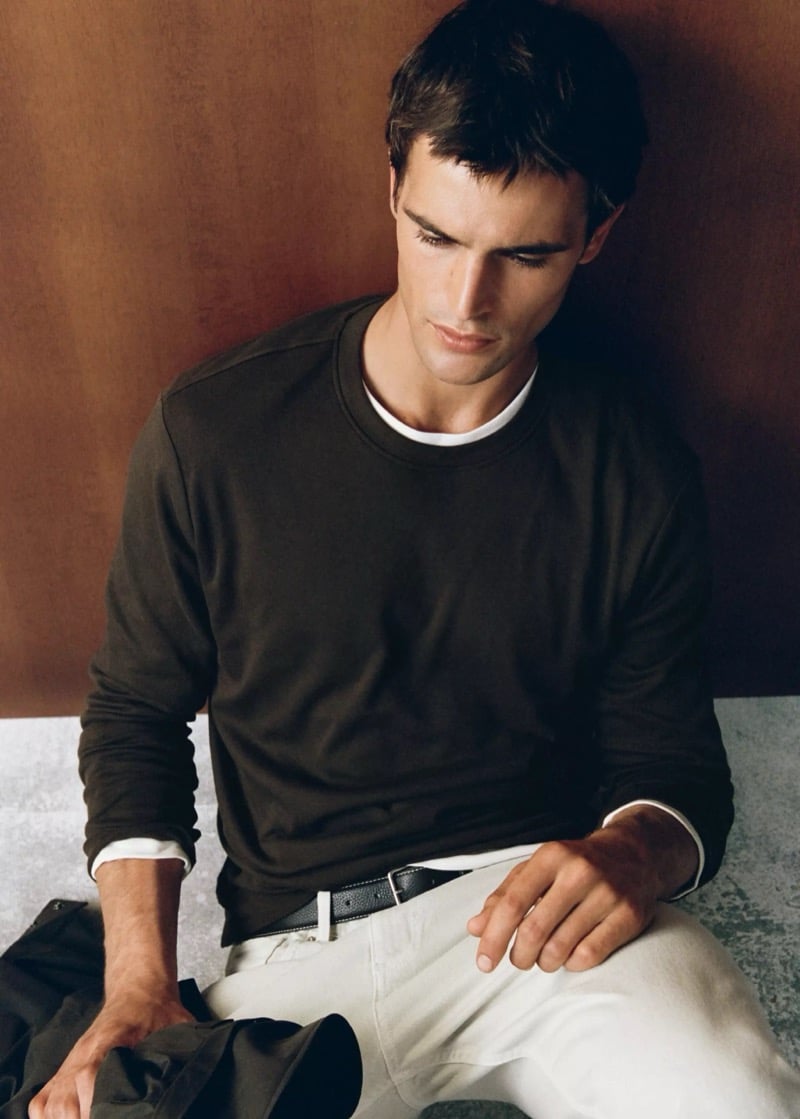 Parker van Noord seated in a dark brown crewneck over a layered tee with light trousers and a black leather belt, a jacket in hand