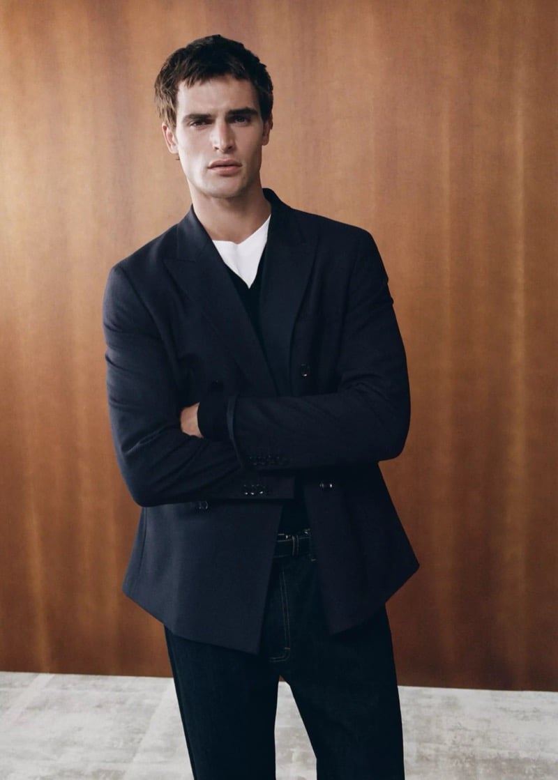 Parker van Noord with arms crossed in a dark double-breasted jacket over a v-neck knit and tee with dark jeans
