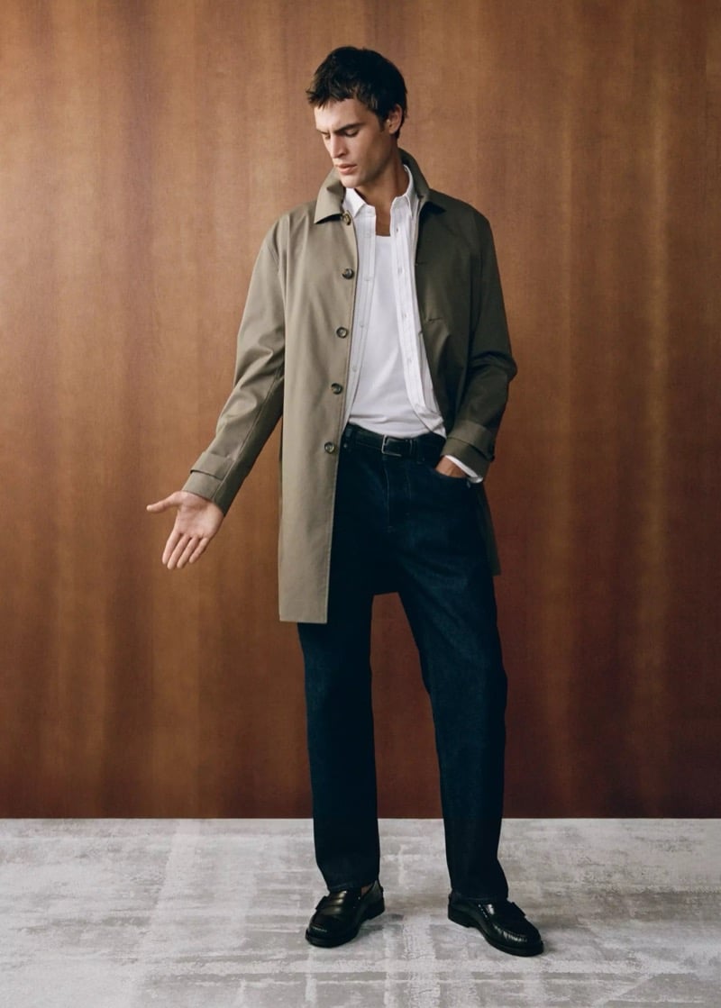 Parker van Noord standing with one hand extended in a button-front gabardine coat over a shirt and tee with dark jeans and loafers