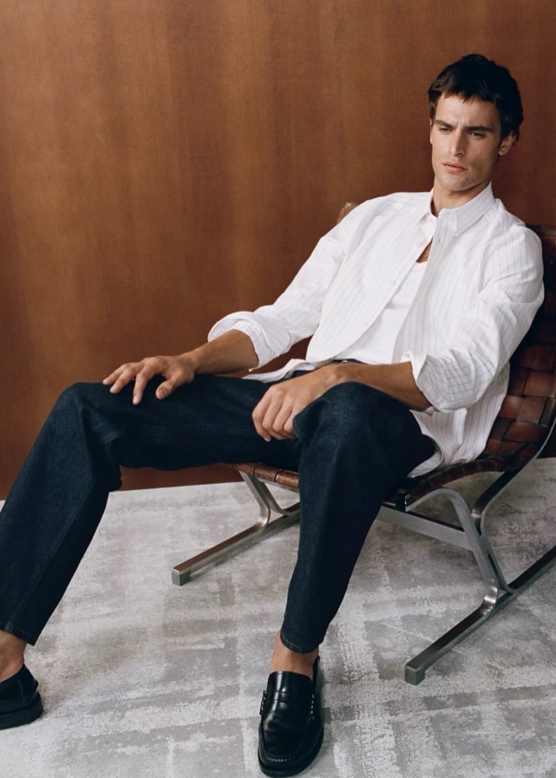 Parker van Noord reclining in a woven leather lounge chair in a pinstripe shirt, dark relaxed-fit jeans, and black loafers