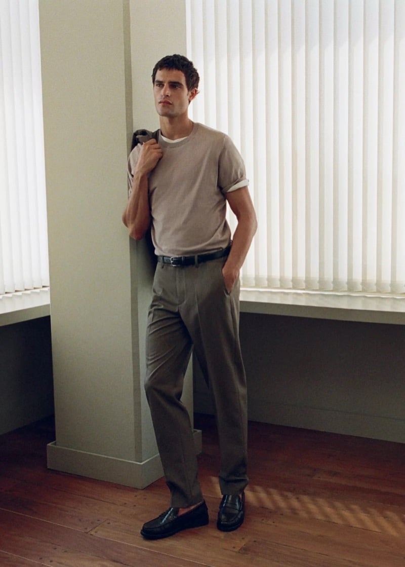 Parker van Noord leaning on a column in a taupe knit tee, pleated olive-gray wool trousers, and black loafers with a jacket over one shoulder