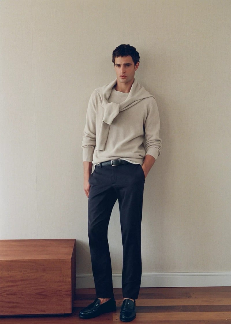 Parker van Noord leaning against a wall in a light knit sweater draped over his shoulders, slim dark chinos, a leather belt, and loafers