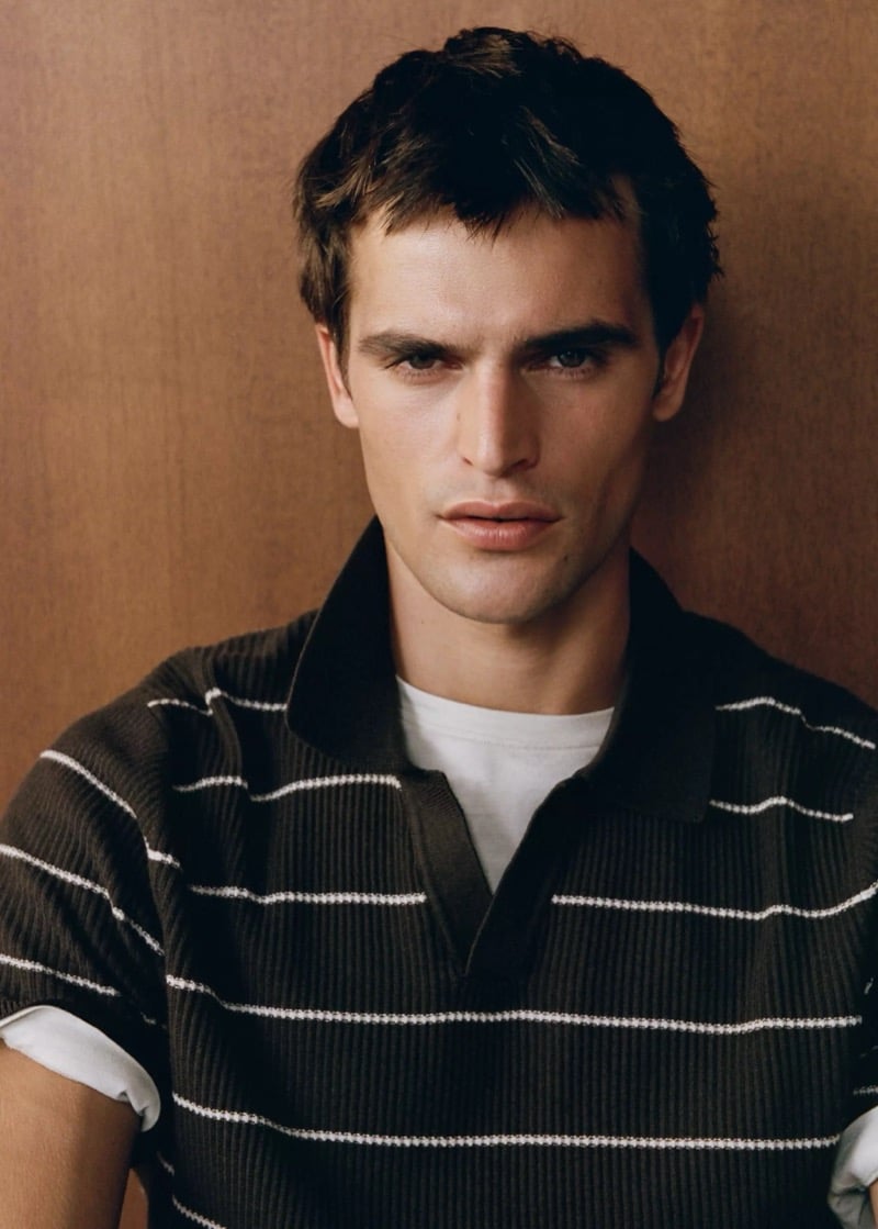 Close-up of Parker van Noord in a dark ribbed knit polo with thin stripes over a tee against a wood-paneled wall