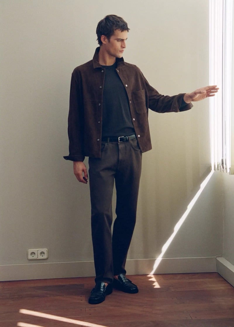 Parker van Noord in a brown suede overshirt, dark tee, and brown jeans reaching toward a sunlit window blind