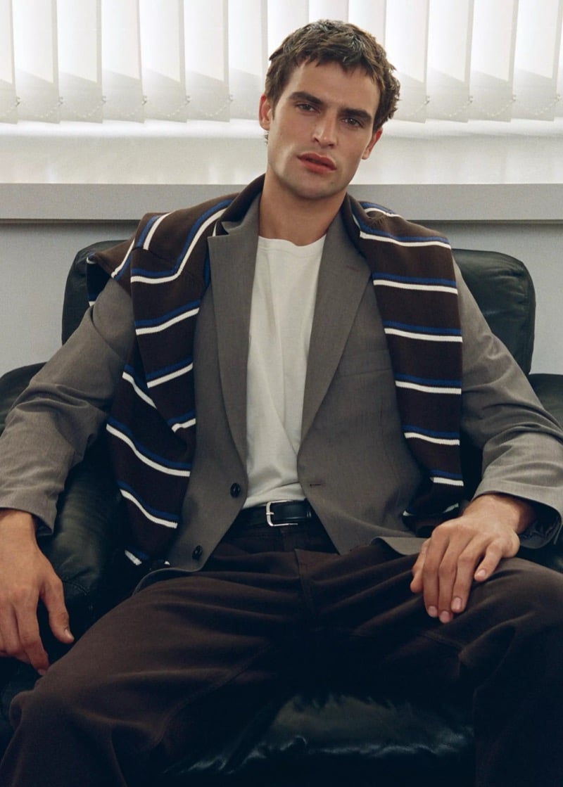 Parker van Noord seated in a black leather armchair in a taupe suit jacket with a striped knit draped over his shoulders and brown trousers