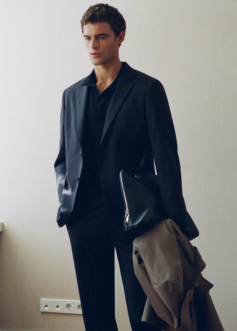 Parker van Noord in a dark wool suit jacket and polo with a zip-top leather portfolio under his arm and a gabardine coat draped over it