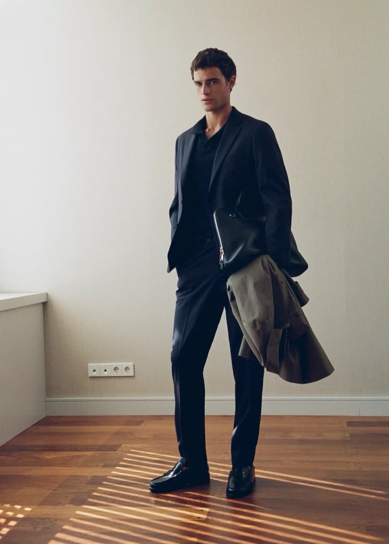 Full-length shot of Parker van Noord in a dark wool suit and polo with loafers, a leather portfolio, and a coat over his arm in a room with striped floor light