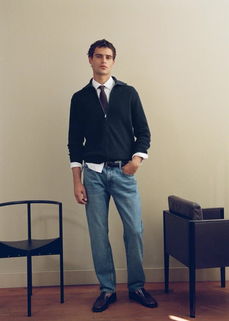 Parker van Noord standing between two armchairs in a dark zip cardigan over a white shirt and tie with light-wash jeans and loafers