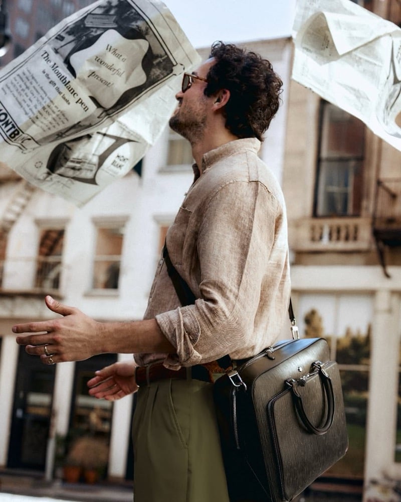Paul Anthony Kelly in profile wearing sunglasses, a linen shirt with rolled sleeves, and olive trousers as newspapers blow past, a Montblanc briefcase at his side