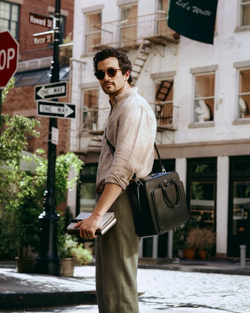 Paul Anthony Kelly in sunglasses and a linen shirt holds notebooks on a New York street corner with the Montblanc Writing Traveler briefcase across his shoulder