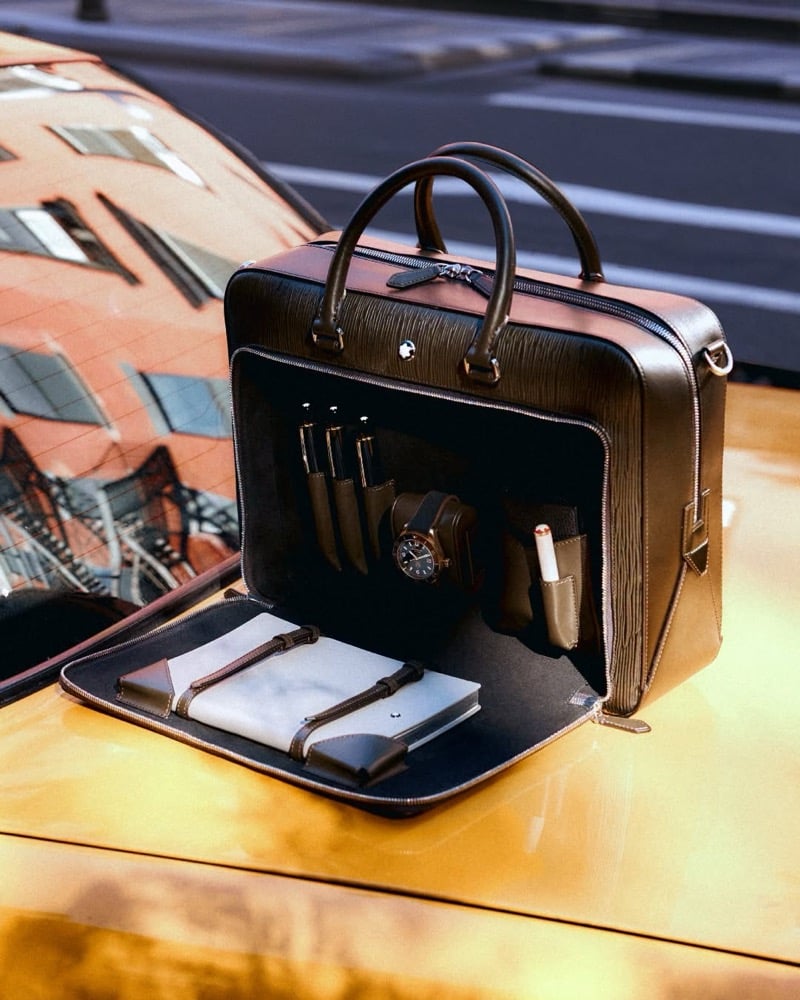 Open Montblanc Writing Traveler briefcase on the hood of a yellow taxi showing pen sleeves, a watch, and a strapped notebook inside