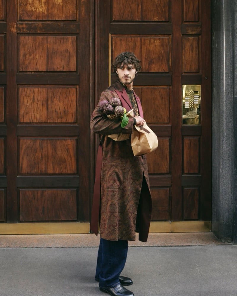 Pedro Pinto stands outside a wood-paneled building entrance holding a paper bag of artichokes, wearing a long burgundy paisley coat over a dark high-neck top with blue jeans and black loafers, for the Etro fall 2026 campaign