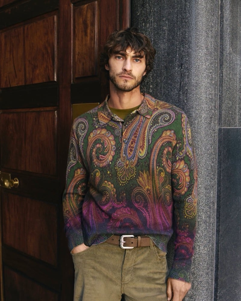 Pedro Pinto leans against a dark wooden door in a green paisley-pattern knit polo that fades to purple and magenta, worn over an olive T-shirt with a brown suede belt and corduroy trousers, for the Etro fall 2026 campaign