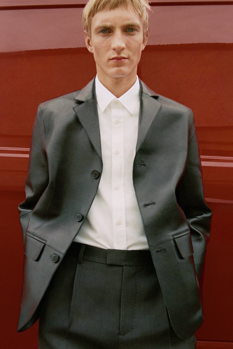 Pierrick Grégoire in a leather blazer, buttoned shirt, and striped trousers in front of a red wall