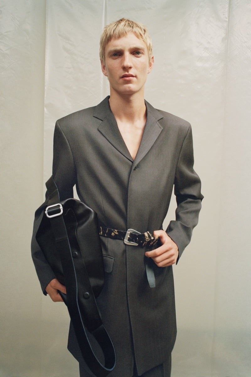 Pierrick Grégoire in a belted long-line suit jacket worn without a shirt, holding a large leather bag