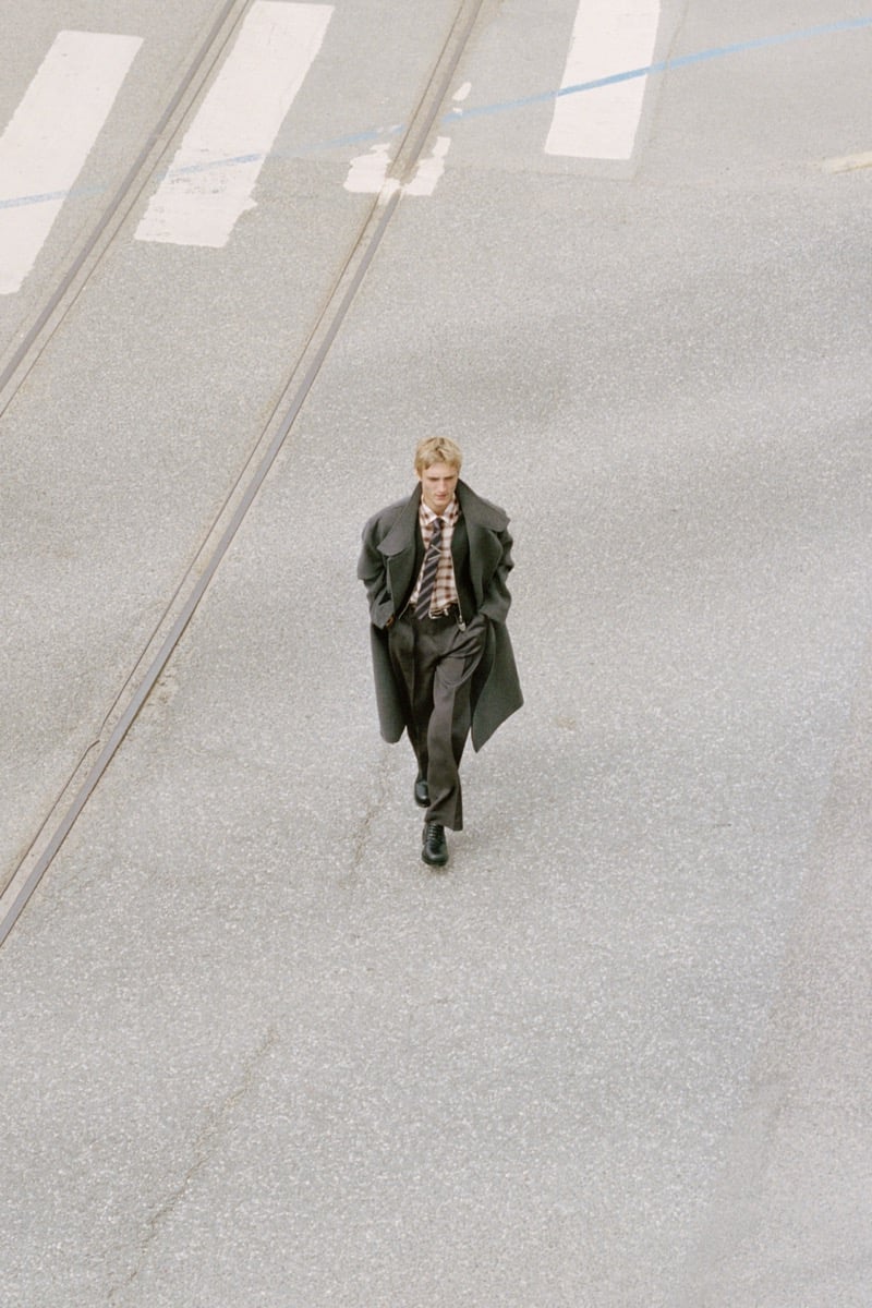 Overhead view of Pierrick Grégoire walking in an oversized coat, plaid shirt, striped tie, and pleated trousers