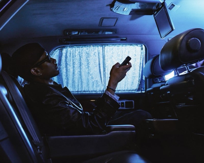 Idriys Ali-Chow reclines in the back seat of a car at night, lit blue, holding a small remote, wearing a leather jacket, cap, and sunglasses beside a curtained window