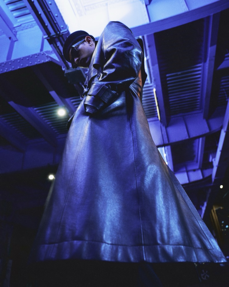 Low-angle shot of Idriys Ali-Chow in a floor-length black leather trench coat, cap, and sunglasses, standing beneath the steel girders of a bridge under blue light