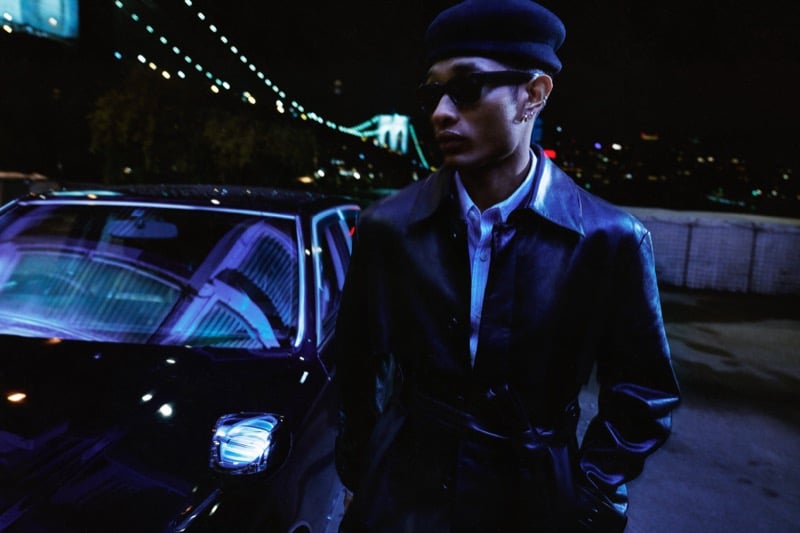 Idriys Ali-Chow stands beside a dark car at night wearing a black leather trench coat, a flat cap, and narrow sunglasses, with a lit suspension bridge and the city skyline blurred behind him