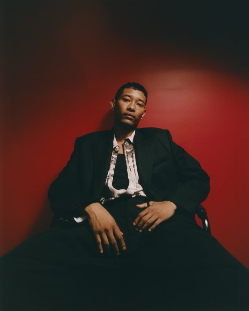Idriys Ali-Chow sits against a deep red backdrop in an oversized dark suit over a printed shirt with a slim tie, hands resting on his knees
