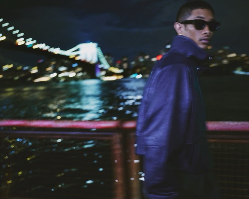 Idriys Ali-Chow turns to look over his shoulder, motion-blurred, on a waterfront walkway at night with a lit suspension bridge and the New York skyline behind him