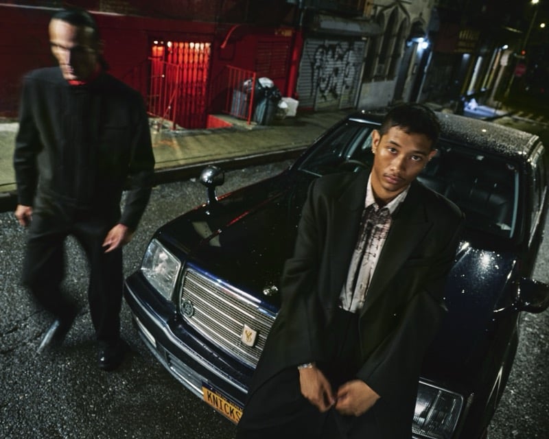 Idriys Ali-Chow leans against a black sedan on a dark New York street lit red from a doorway, wearing a dark suit over a marbled-print shirt, with a blurred figure of Scott Barnhill walking behind in dark clothing