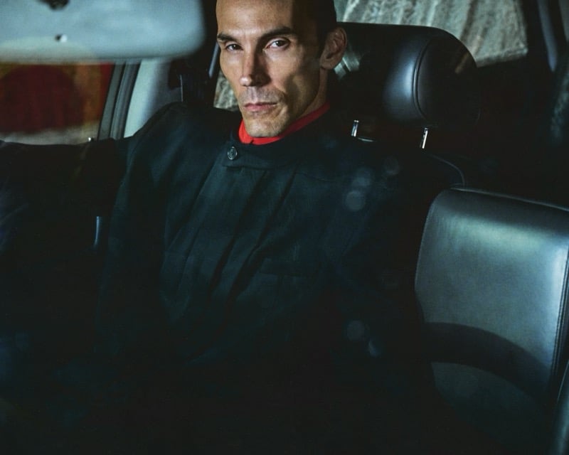 Scott Barnhill sits in the back seat of a car at night, lit blue, wearing a dark mandarin-collar jacket over a red stand collar