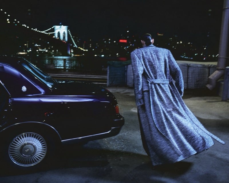Scott Barnhill walks away from the camera toward the waterfront in a long marled wool overcoat with a belted back, a dark car beside him and a lit bridge in the distance