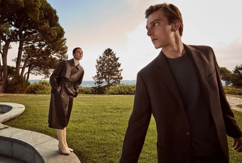 Quentin Demeester in a soft blazer over a crewneck on a villa lawn with Beauïse Genç behind him in a belted leather trench coat