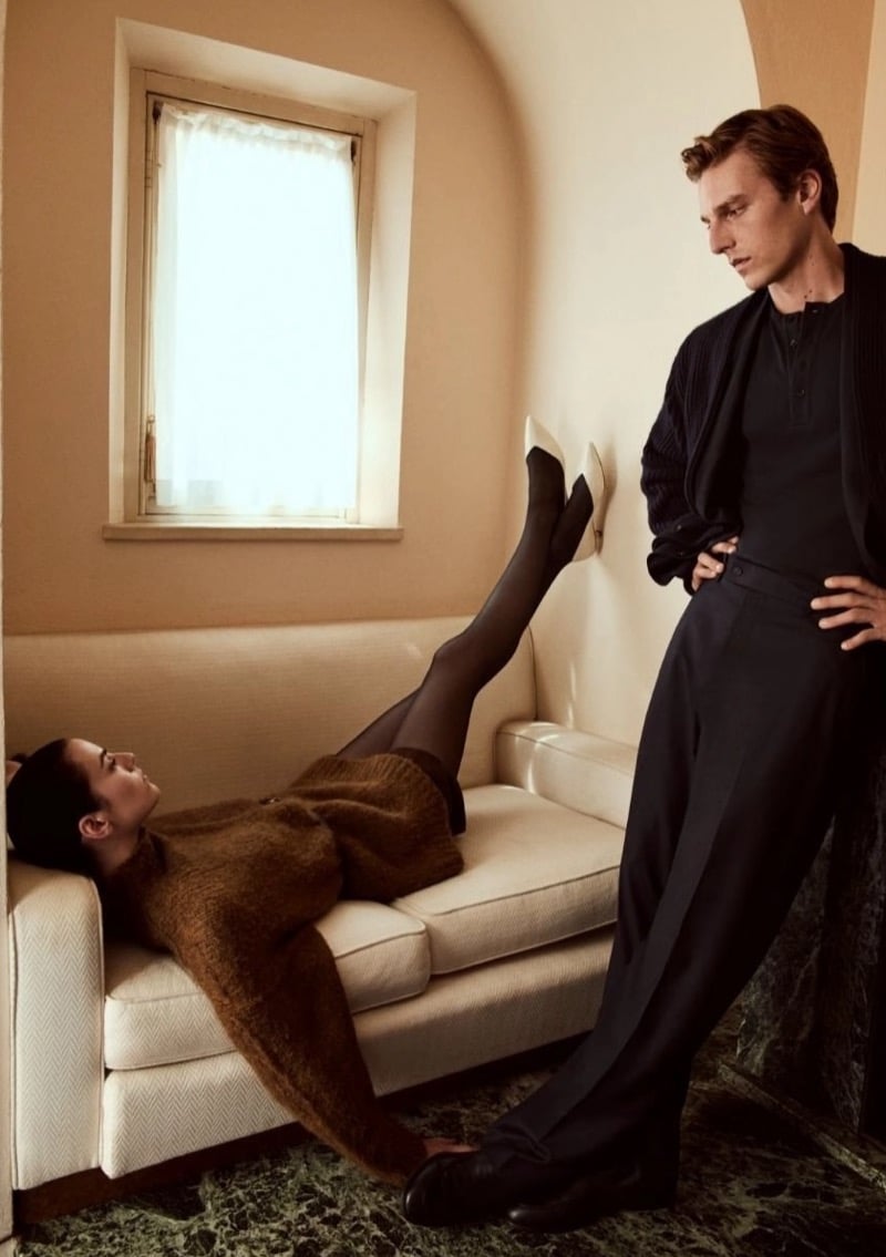 Beauïse Genç lying on a sofa with her legs raised against the wall as Quentin Demeester stands nearby in a cardigan, henley, and high-waisted trousers