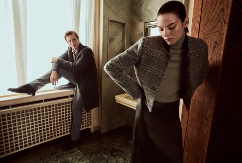 Quentin Demeester perched on a windowsill in an overcoat as Beauïse Genç stands in front in a plaid cropped jacket, turtleneck, and leather skirt
