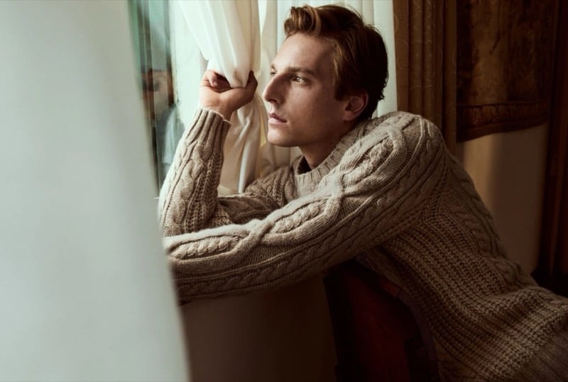 Quentin Demeester leaning on his arms at a curtained window in a chunky cable knit sweater
