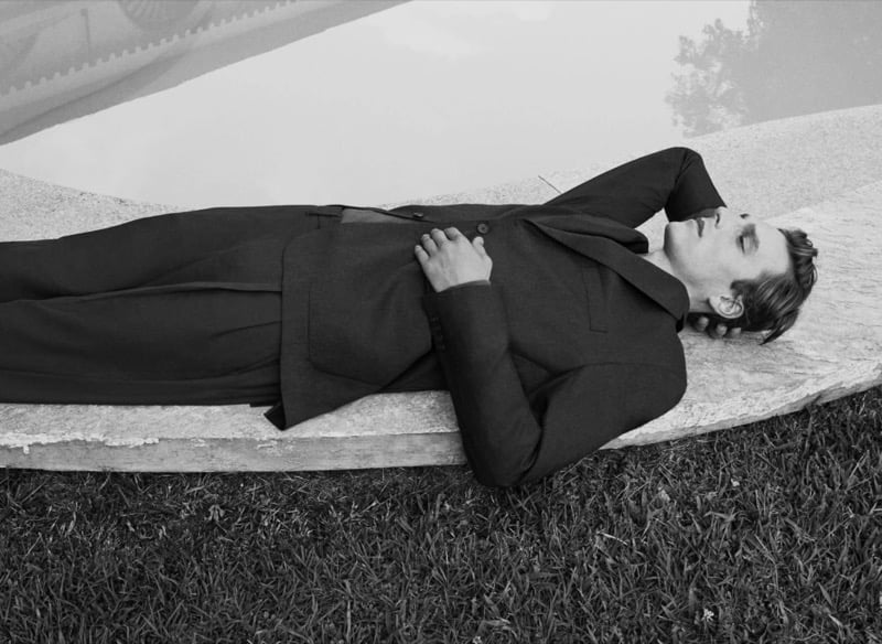 Black-and-white image of Quentin Demeester lying on a stone ledge beside a pool in a tailored jacket and trousers