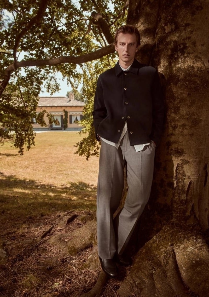 Quentin Demeester leaning against a tree trunk in a button-front jacket, open-collar shirt, and gray wide-leg trousers, with a villa in the distance