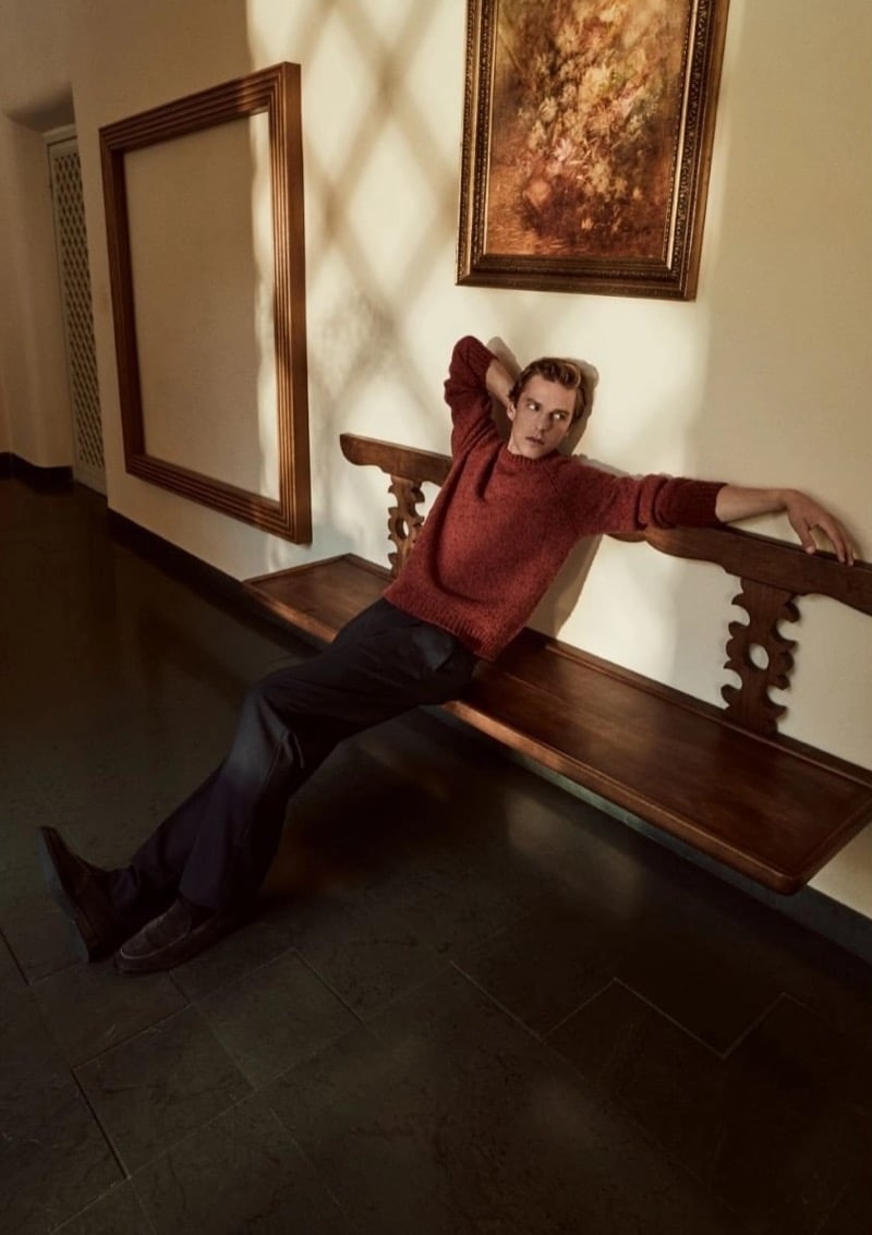 Quentin Demeester stretched across a carved wooden bench below a gilt-framed painting, wearing a knit crewneck sweater, relaxed trousers, and loafers