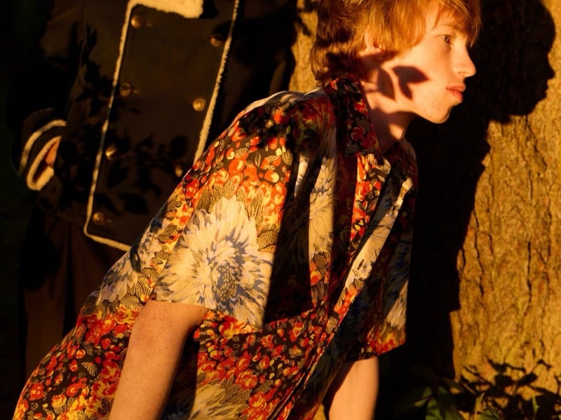 Cullan Sturdevant leans forward in a floral-print Dior shirt beside a tree while another model in a shearling-trimmed coat stands behind
