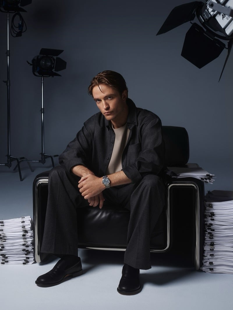 Robert Pattinson seated on a leather armchair between stacks of clipped papers, with studio lights overhead, wearing an overshirt, wide trousers, and a steel Jaeger-LeCoultre watch
